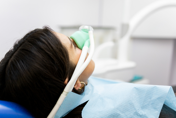 An adult woman sits in a dentist's chair wearing a nasal mask to inhale nitrous oxide.