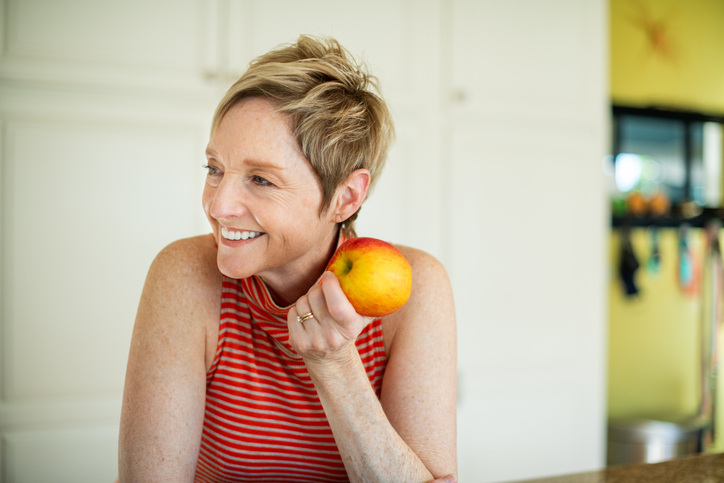 Stylish woman with a great smile with dental implants leans on her kitchen counter while eating an apple.