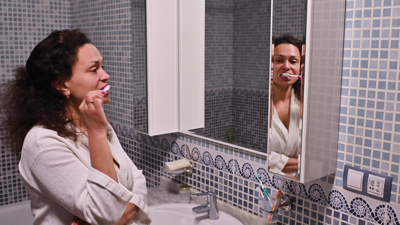 A woman brushing her teeth in a tiled bathroom while looking into the mirror.